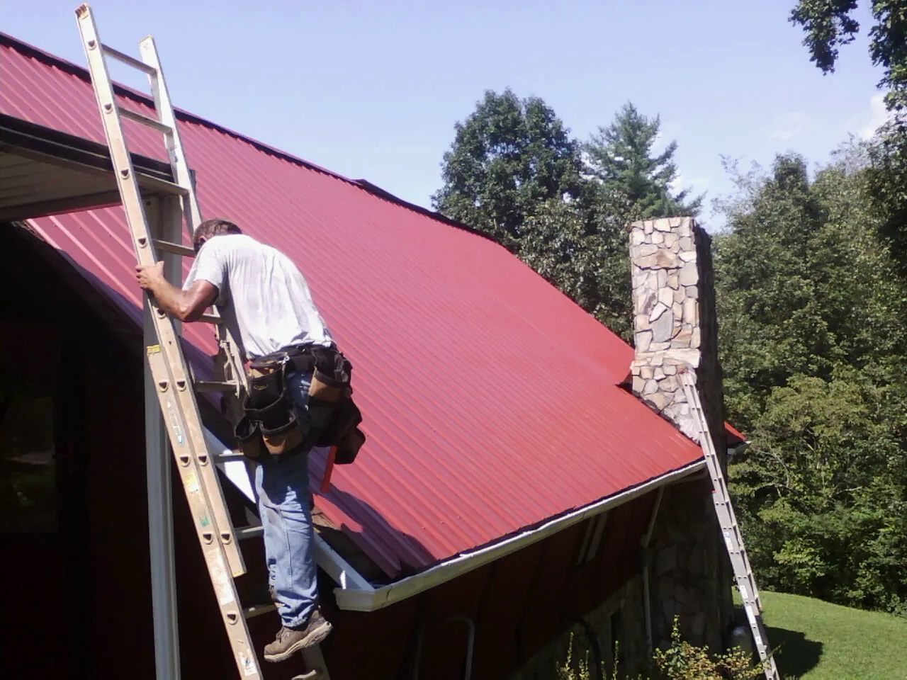 Roofer on a ladder beside a red metal roof and stone chimney — Soffit & Fascia Repair by Roofing Brothers in Enon Valley